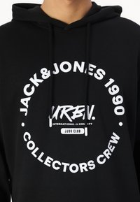 Schwarzer Kapuzenpullover mit weißem Drucktext und kreisförmigen Logos. Der Stoff ist weich und verfügt über eine Kapuze sowie verstellbare Kordeln.