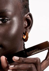 MAC Studio Radiance 24HR Luminous Lift Concealer i ett mörkt rör med guldtext; modell som håller den med en brun hudton och glänsande örhängen.
