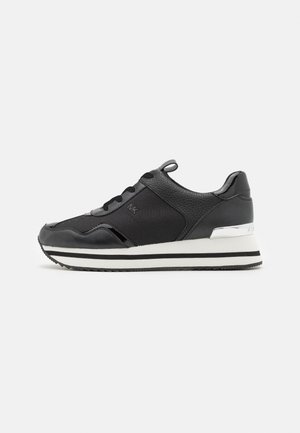 Baskets de sport noires avec une tige synthétique texturée, ornées d'un bord lisse, d'accents argentés et de rayures blanches sur une semelle noire.