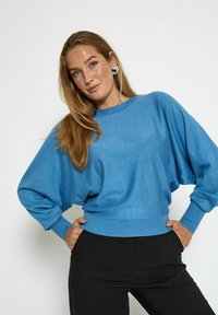 Pullover oversize blu chiaro con collo rotondo, polsini e orlo a coste. Abbinato a pantaloni neri aderenti, che mostrano un design rilassato.