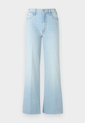 Mladá žena stojaca so založenými rukami, oblečená v čiernom tielku bez rukávov, svetlomodrých širokých džínsoch a čiernych sandáloch na vysokom opätku na sivom podlahe.