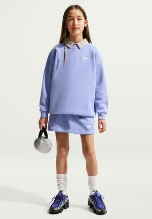 Ragazza con felpa e gonna Nike viola chiaro, che tiene una piccola borsa rotonda d'argento, con calzini bianchi e scarpe da ginnastica viola.