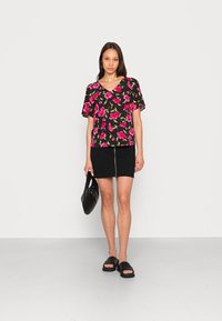 Blusa preta com padrão floral, com flores rosas, decote em V, mangas curtas, acompanhada de uma saia lápis preta com fecho à frente, segurando uma bolsa preta.