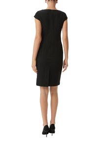 Schwarzes, eng anliegendes Kleid mit kurzen Ärmeln, Rückenzipper und vertikalen Nähten; knielang mit einem Schlitz hinten. Getragen mit schwarzen High Heels.