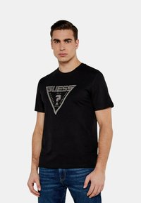 T-shirt in cotone nero con un logo triangolare silver che presenta la scritta "GUESS" e un punto interrogativo. Collo rotondo e maniche corte.