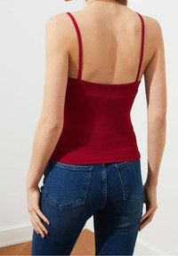 Červený žebrovaný top na tenké ramínka v kombinaci s tmavě modrými džíny. Má přiléhavý střih, hladkou texturu a viditelné švy na zadní straně.