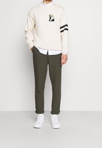 Krämfärgad stickad tröja med en grafisk applikation, svarta randiga detaljer på ärmarna, olivgröna byxor och vita sneakers.