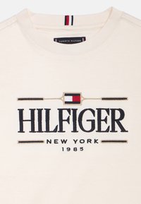 T-shirt en coton couleur crème avec le logo "HILFIGER" brodé en noir, accompagné de détails en rouge, blanc et bleu.