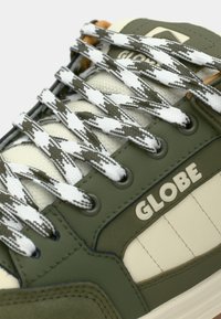 Gros plan sur une chaussure de sport verte et beige avec des lacets blancs et gris à motif et le logo "GLOBE" sur le panneau latéral.