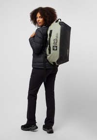 Green and black duffel bag with adjustable straps, featuring a logo and a sturdy design. Person wearing a black puffer jacket and black pants.