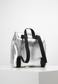 Sac à dos métallique argenté à la forme structurée. Comprend des sangles et des accents noirs. Texture lisse avec une finition brillante.