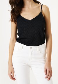 Černá texturovaná tílko s výstřihem do V a úzkými ramínky, kombinováno s bílými džíny. Látka má jemný vzor a lehký design.