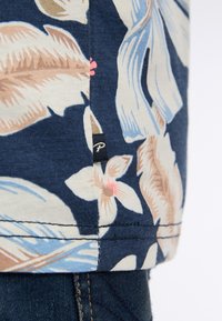 Yksi kukkakuosisista yläosista, jossa on tummansininen pohja ja kermaisia, sinisiä ja vaaleanpunaisia sävyjä. Lippulappu on pieni ja musta helman kohdalla, ja ompeleet ovat näkyviä.