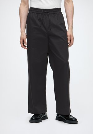 Mann trägt lockere schwarze Hose mit elastischem Bund und Bindeband, kombiniert mit schwarzen Lederschuhen und weißem Hemd.