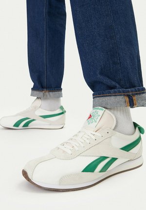 Zapatillas Reebok blancas y verdes usadas con calcetines blancos y jeans azules remangados sobre un fondo blanco.