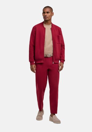 Mann trägt rote Reißverschlussjacke und passende Hose, beige Hemd und Gürtel sowie beige Slipper und steht vor weißem Hintergrund.