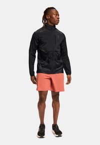 Schwarze leichte Jacke mit Reißverschluss, kontrastierenden helleren Einsätzen, langen Ärmeln, kombiniert mit korallenfarbener Shorts und schwarzen Sportschuhen.