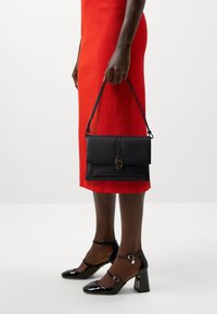 Borsa a tracolla nera in pelle texture con pattina frontale e chiusura in hardware dorato, abbinata a décolleté in pelle verniciata nera e un vestito rosso.