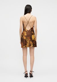 Γυναίκα που φοράει καφέ μίνι φόρεμα με floral σχέδιο και λεπτούς, σταυρωτούς ιμάντες στην πλάτη, και μαύρα πέδιλα με ψηλό τακούνι, στέκεται μπροστά σε απλό φόντο.