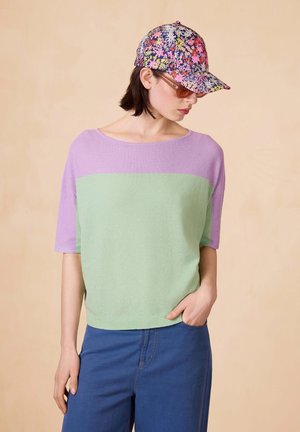Korte mouwtrui in gestructureerd groen en lavendel, gecombineerd met een bloemenpet. Het model draagt blauwe denim broek en poseert tegen een neutrale achtergrond.