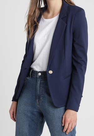 Žena v námořnicky modrém saku přes bílé tričko, v kombinaci s modrými džíny s vysokým pasem, stojící před jednobarevným pozadím.