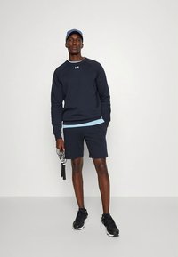 Marineblauw sportief sweatshirt met lichtblauwe accent, gecombineerd met marineblauwe korte broek. Model houdt een springtouw vast, draagt zwarte sportschoenen en een pet.