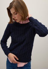 Dunkelblauer gestrickter Pullover mit einem Zopf-Muster und gerippten Bündchen, kombiniert mit einem Kragenhemd mit weißen und roten Streifen.