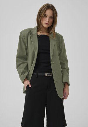 Frau trägt einen übergroßen olivgrünen Blazer, schwarzes trägerloses Oberteil, schwarze weit geschnittene Hosen und einen schwarzen Gürtel mit silberner Schnalle vor einfarbigem Hintergrund.