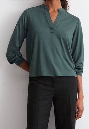 Vrouw draagt een teal langärmelig V-hals top met gerimpelde mouwen en zwarte op maat gemaakte broek, tegen een witte achtergrond.