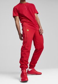 Persona che indossa pantaloni e t-shirt con marchio Ferrari rossi, con sneakers Puma rosse, in piedi su uno sfondo grigio uniforme.