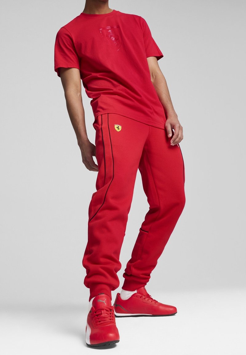Persona che indossa pantaloni e t-shirt con marchio Ferrari rossi, con sneakers Puma rosse, in piedi su uno sfondo grigio uniforme.