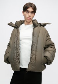 Casaco acolchoado em verde oliva, com capuz, fecho éclair e punhos canelados. O tecido possui uma textura suave com padrões acolchoados.