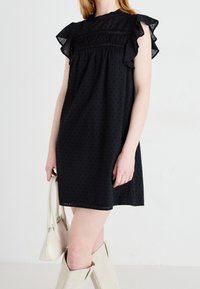 Musta mekko, jossa on lyhyet röyhelöhihoja, korkea kaula-aukko ja rypytetty yksityiskohta vartalon kohdalla, valmistettu tekstuurisesta kankaasta.