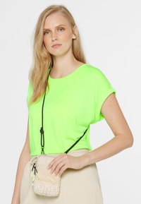 Neon-grünes T-Shirt mit kurzen Ärmeln und rundem Ausschnitt. Ein schwarzer verstellbarer Riemen hält eine kleine beigefarbene Umhängetasche mit weißem Text.