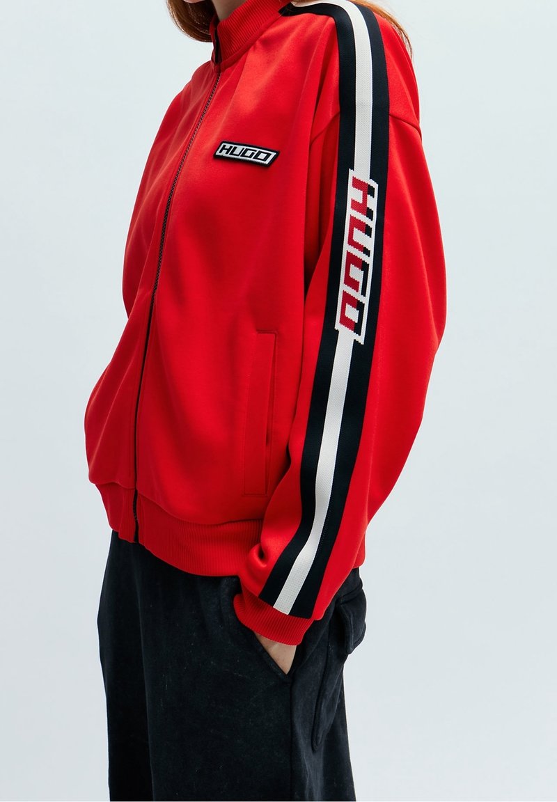 Červená zapínací bunda s vysokým límcem, s černobílými pruhy na rukávech a s nápisem "HUGO" na přední části a na rukávu.