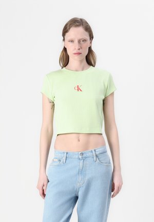 Vaaleanvihreä lyhytnenäinen T-paita ribbitekstuurilla, jossa on pieni punainen CK-logo keskellä. Yhdistetty löysiin sinisiin farkkuihin.
