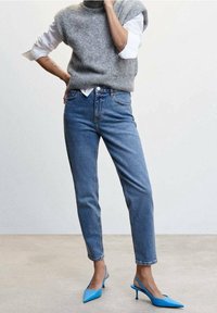 Szary, dziany sweter bez rękawów noszony na białej koszuli, w zestawie z niebieskimi spodniami typu high-waist i niebieskimi szpilkami z czubkiem.