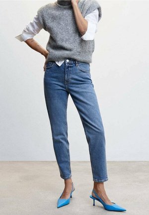 Suéter gris de punto sin mangas sobre una camisa blanca, combinado con pantalones vaqueros de tiro alto azules y tacones de punta azul.