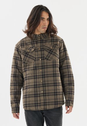 Man met lang haar die een bruin-zwart geruit overhemd draagt met twee borstzakken aan de voorkant, staand tegen een witte achtergrond.