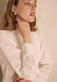 Blusa bianca con texture, polsini arricciati e dettagli sottili di ricamo, abbinata a grandi orecchini dorati. Tessuto liscio e leggero.