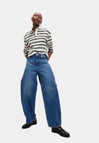 Gestreept overhemd met lange mouwen in crème en zwart, gecombineerd met wijde blauwe jeans. Model draagt zwarte instappers, wat een ontspannen uitstraling toont.