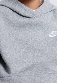 Šedá bavlněná mikina s klokaní kapsou, kapucí na šňůrku a bílým logem Nike. Látka má měkký, strukturovaný povrch.