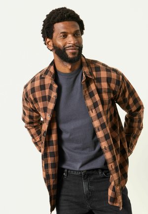 Man with curly hair and beard wearing brown and black checkered shirt open over a dark gray t-shirt, with hands behind his back.