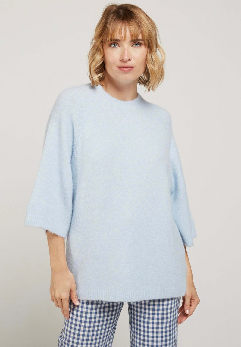 Femme aux cheveux blonds courts portant un pull en peluche bleu clair à manches jusqu'au coude et un pantalon à carreaux bleu et blanc, se tenant devant un arrière-plan uni.