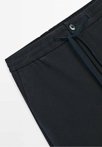 Pantalon en tissu noir avec taille élastique, fermeture par bouton et cordon noué visible sur fond blanc.