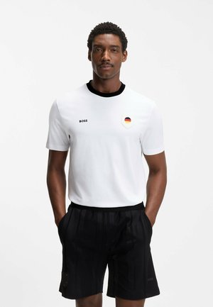 Mężczyzna w białej koszulce z czarnym kołnierzem i naszywką z flagą Niemiec, w czarnych spodenkach, stoi z rękami w kieszeniach.