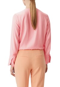 Rosa Bluse mit langen Ärmeln, abgerundetem Halsausschnitt und dezenten seitlichen Schlitzen an den Manschetten, kombiniert mit pfirsichfarbenen, taillierten Hosen.