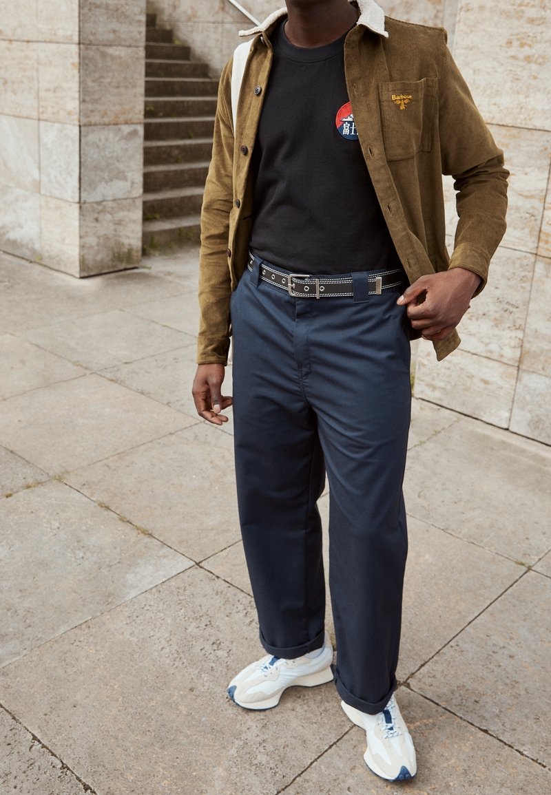 Homme portant une veste marron, un t-shirt noir à motif graphique, un pantalon bleu marine avec une ceinture noire, et des baskets blanches, debout sur un pavé en pierre près d'un escalier.