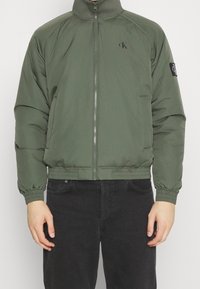 Olijfgroene gewatteerde jas met een hoge kraag, een ritssluiting aan de voorkant, elastische manchetten en tailleband. Heeft een logopatch op de linkermouw.