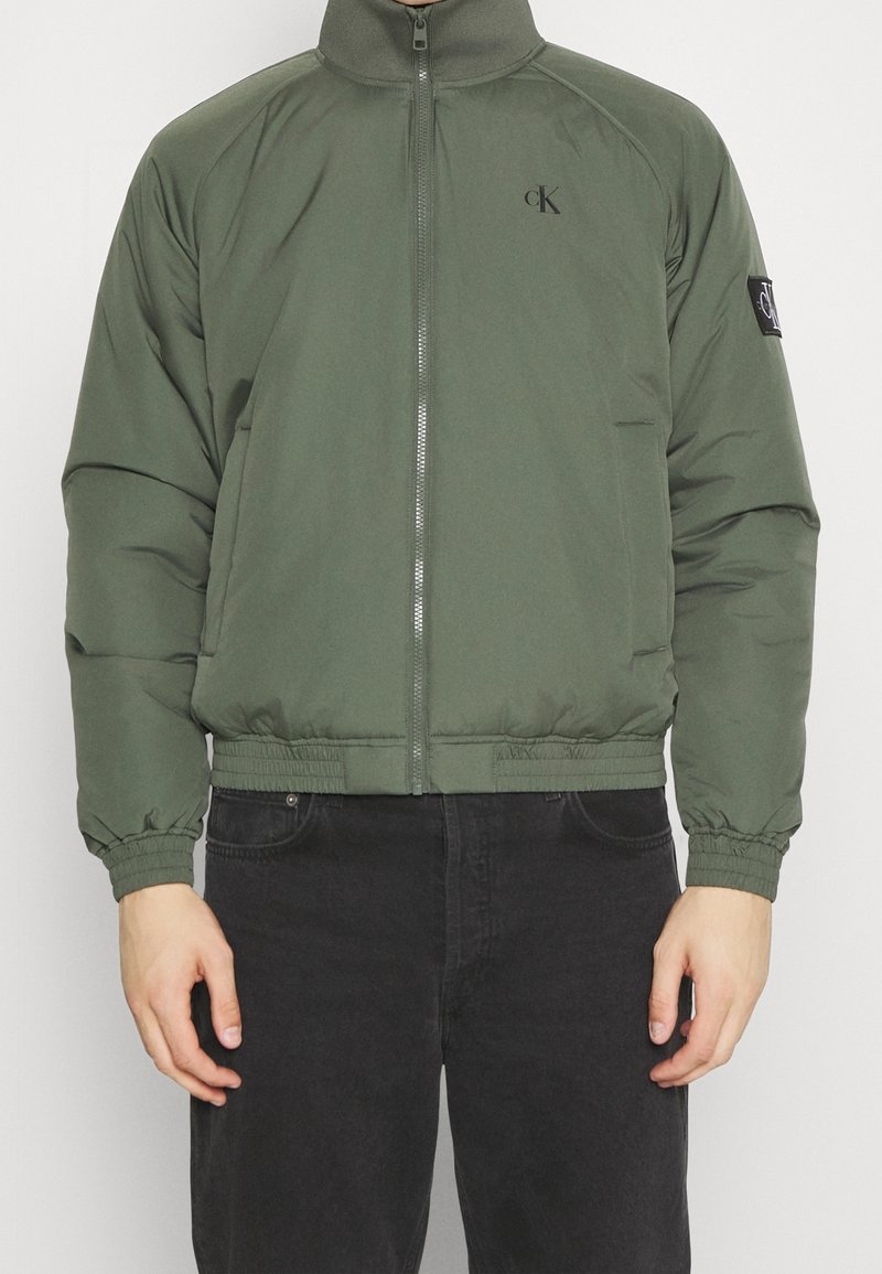 Olijfgroene gewatteerde jas met een hoge kraag, een ritssluiting aan de voorkant, elastische manchetten en tailleband. Heeft een logopatch op de linkermouw.